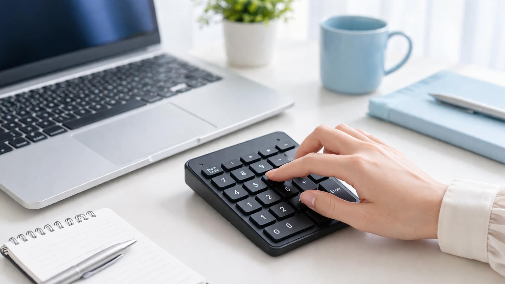 ノートパソコンの横に置いた外付けテンキーを、女性の手で操作している実写風のデスク写真